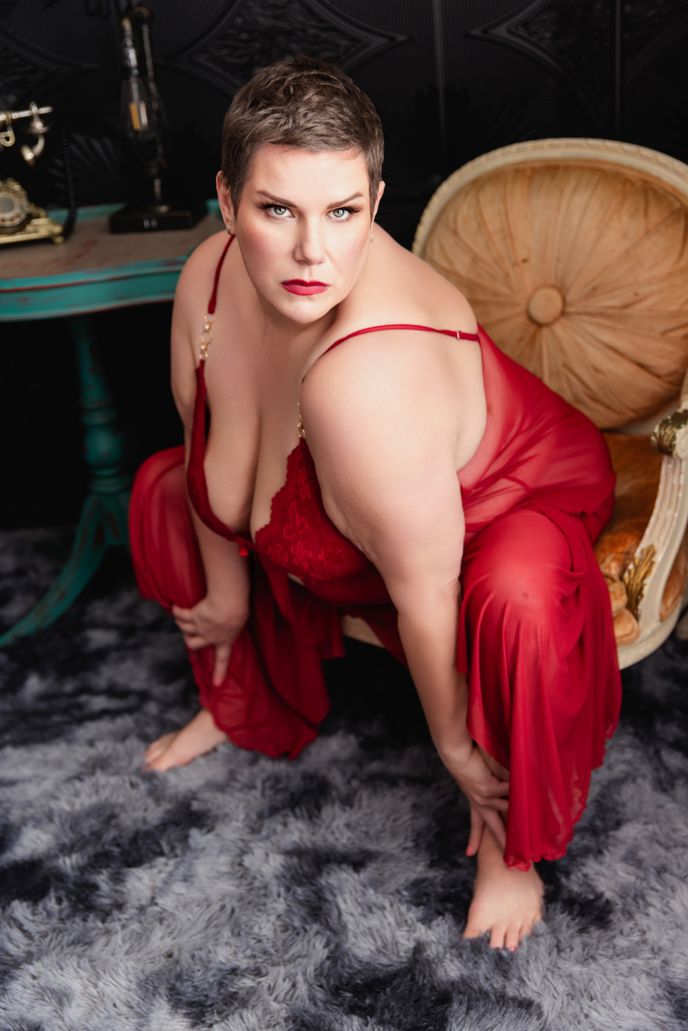 A woman wearing a red slip sits in a chair while holding her ankles in a studio after visiting makeup artists in indianapolis