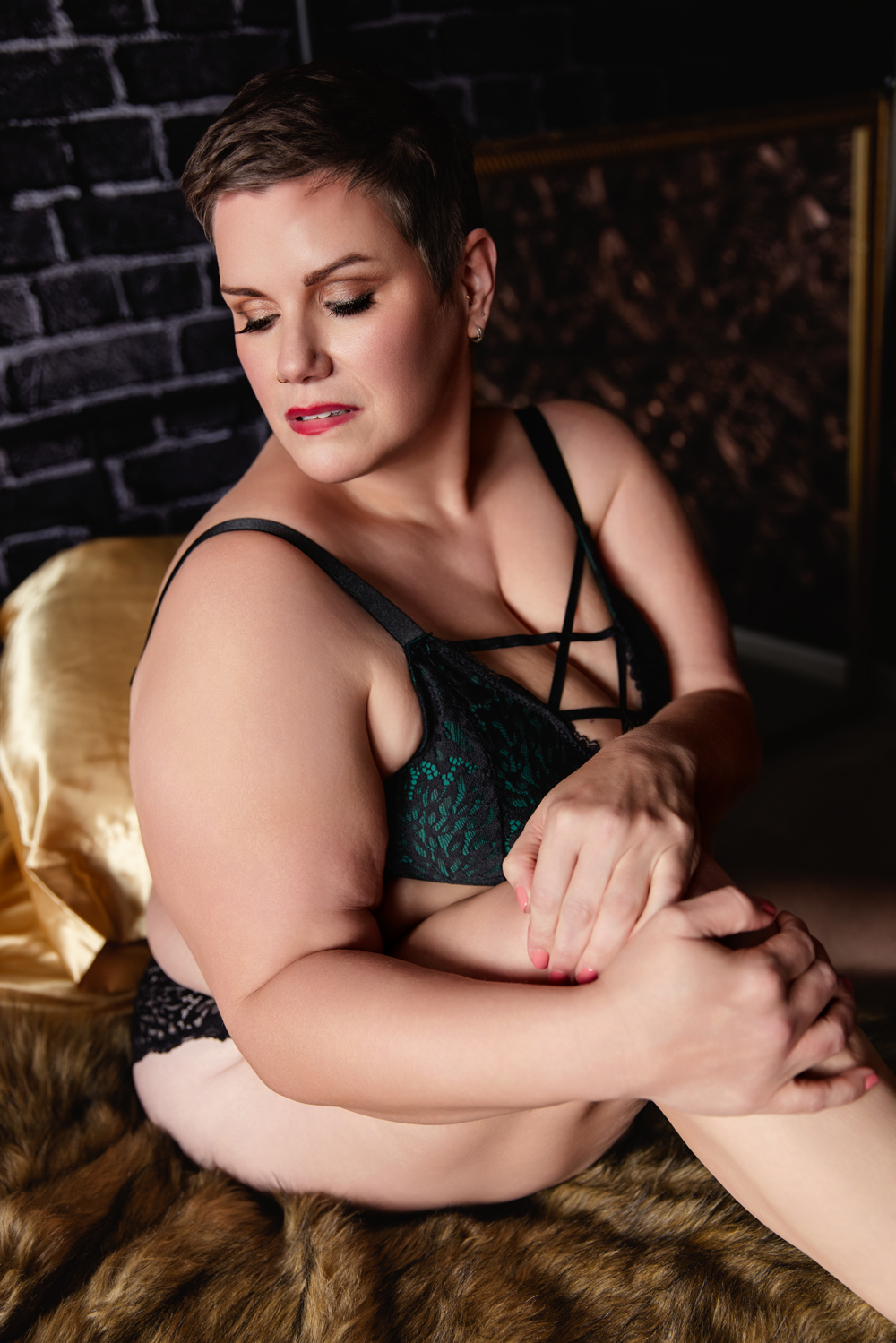 A woman in green lingerie sits across a bed with hands on her knee in a studio after visiting makeup artists in indianapolis