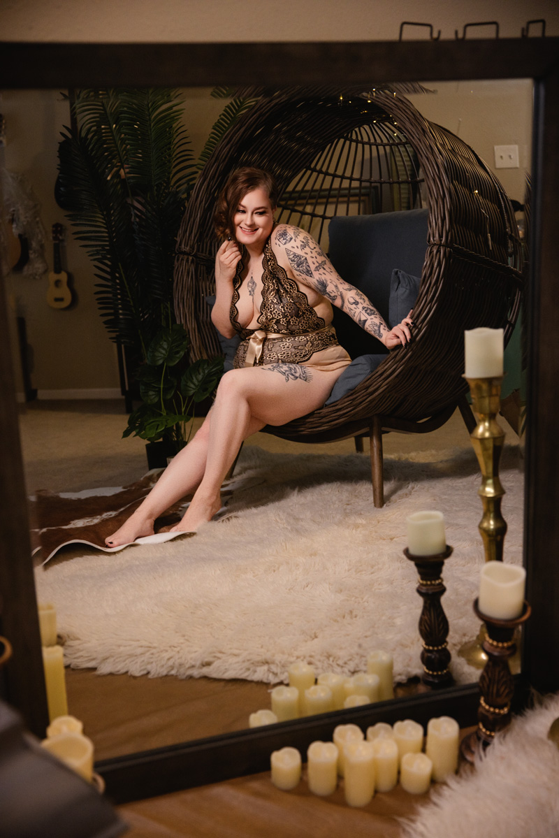 A tattooed woman giggles while sitting in a large dark wicker egg chair in a studio in black lace lingerie after visiting nail salons in indianapolis