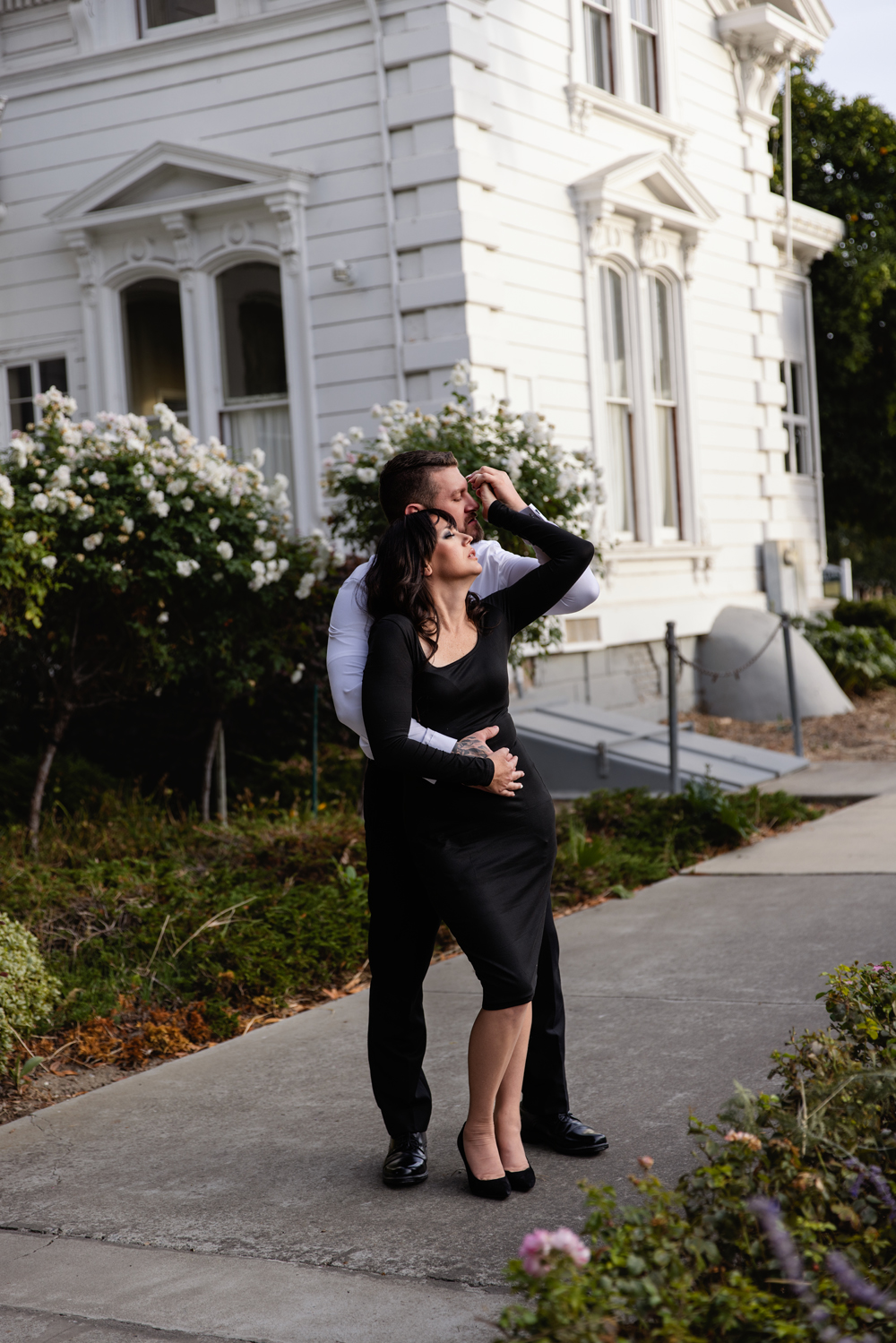 A man and woman snuggle while holding hands on a sidewalk in a black dress and white shirt