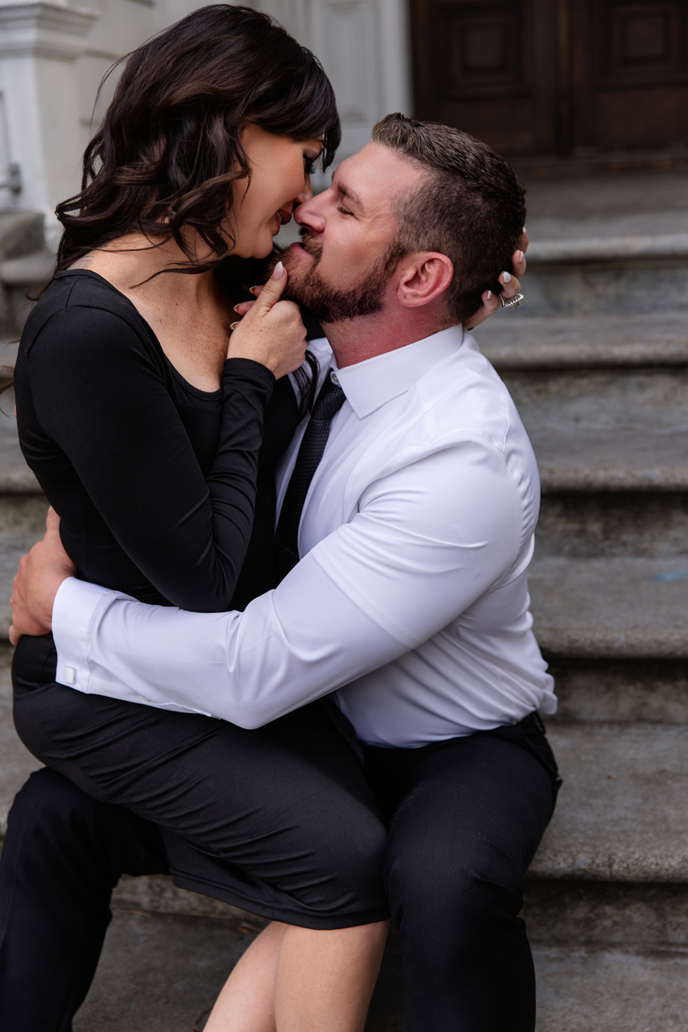 A happy couple lean in for a kiss while she sits on his lap on a set of stairs after visiting romantic restaurants in indianapolis