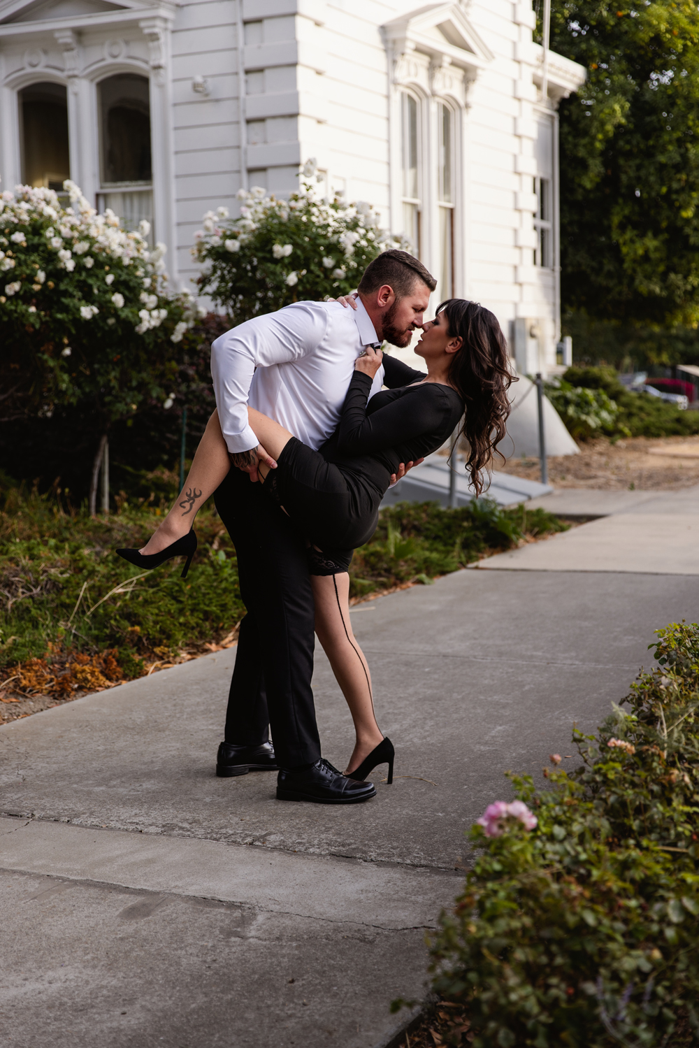 A man in a white shirt leans in for a kiss with his wife in a black dress on a sidewalk while holding her leg up after visiting romantic restaurants in indianapolis